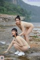 A couple of naked women sitting on top of a rock next to a river.