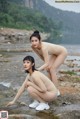 A couple of naked women sitting on top of a rock next to a river.