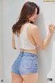 A woman in a white top and blue denim shorts leaning against a wall.