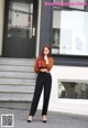 A woman standing in front of a building wearing a brown sweater and black pants.